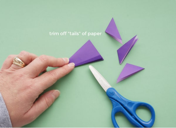 folded paper snowflake with trimmed off tails of paper and a pair of scissors and hand holding the folded paper