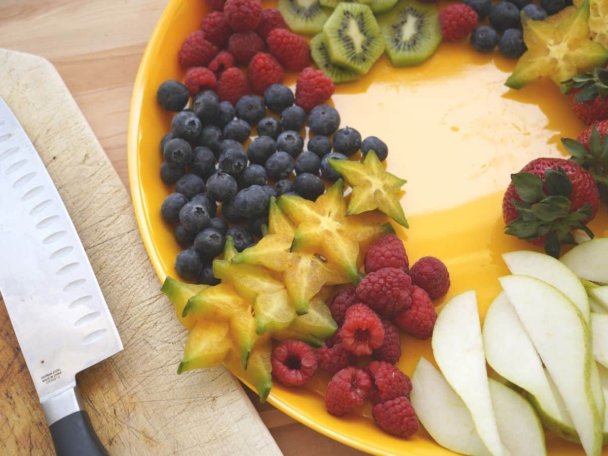 sliced starfruit kiwi blueberries raspberries and pear slices on yellow tray with knife on cutting board