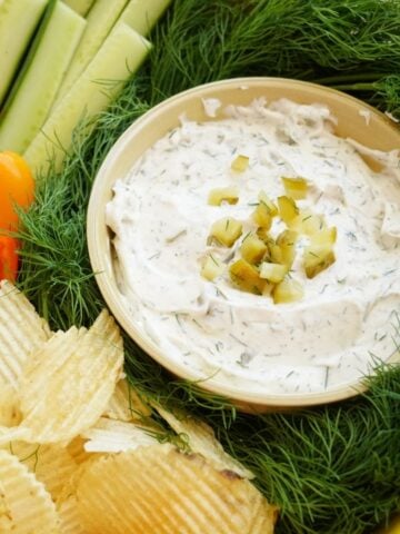 close up of bowl of dill pickle dip surrounded by chips and vegetables and fresh dill