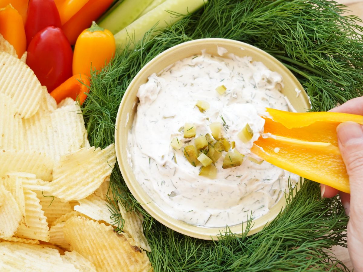 yellow pepper dipped into bowl of dill pickle dip surrounded by chips and cucumbers