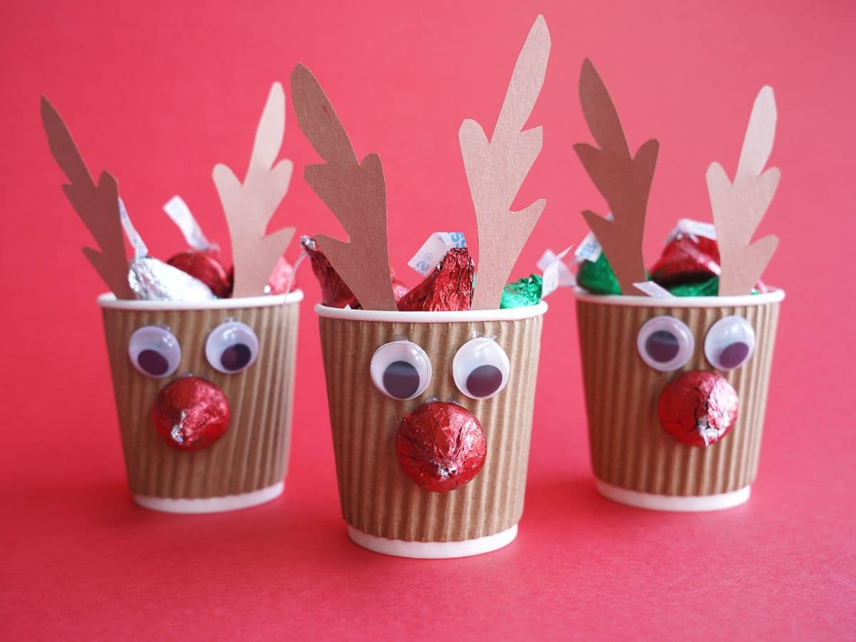 3 reindeer cups filled with chocolate kisses lined up with red background