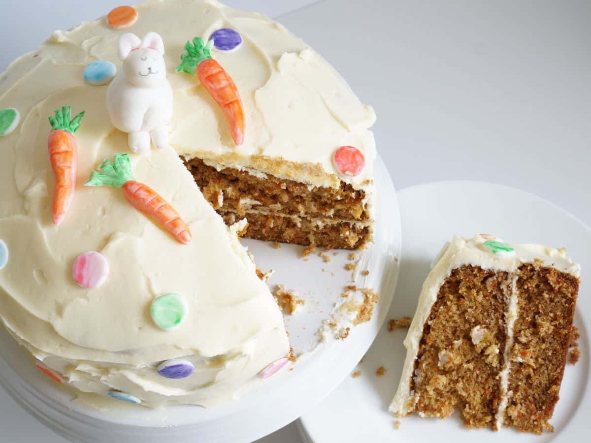 carrot cake with cream cheese frosting with slice cut out of the cake and fondant bunny and carrots on top