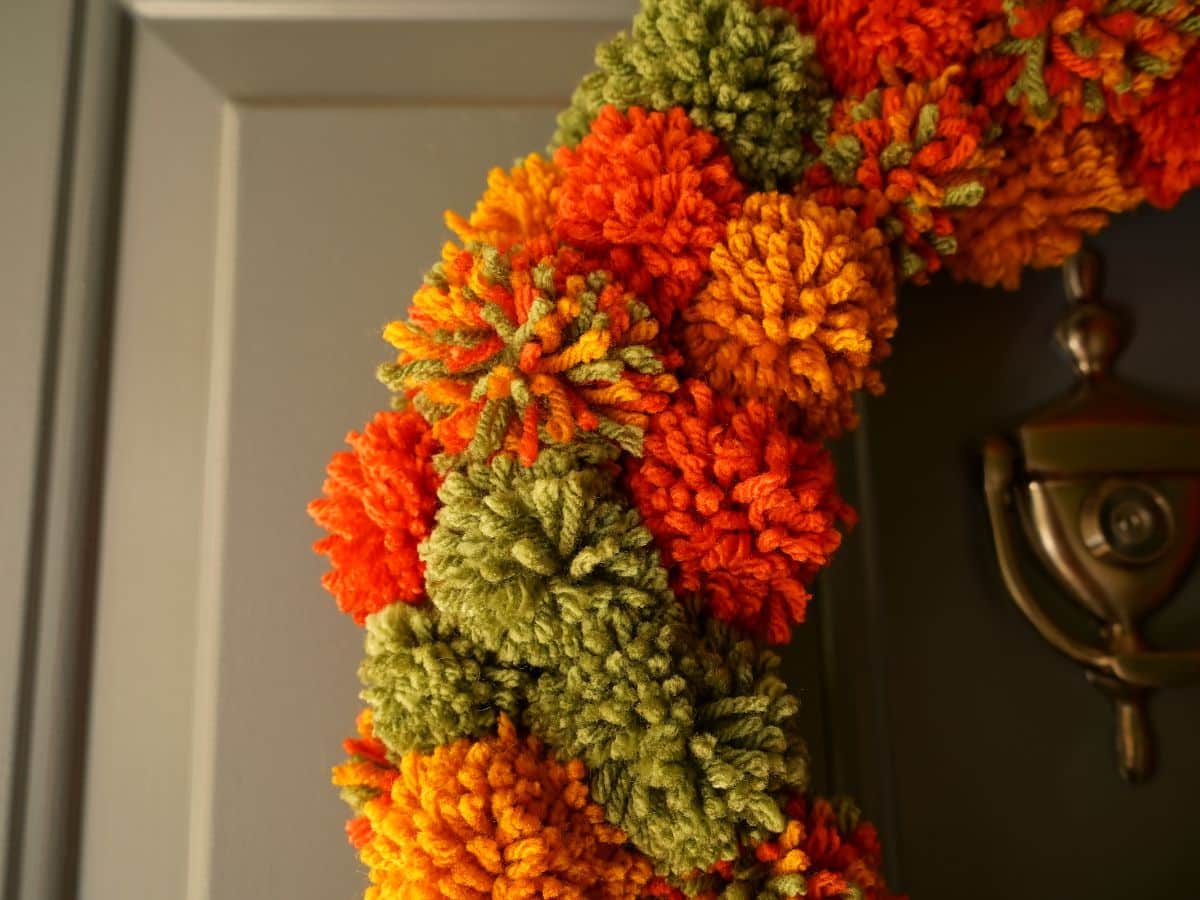 close up of fall pompom wreath with green, orange, and dark yellow pompoms hanging on a door