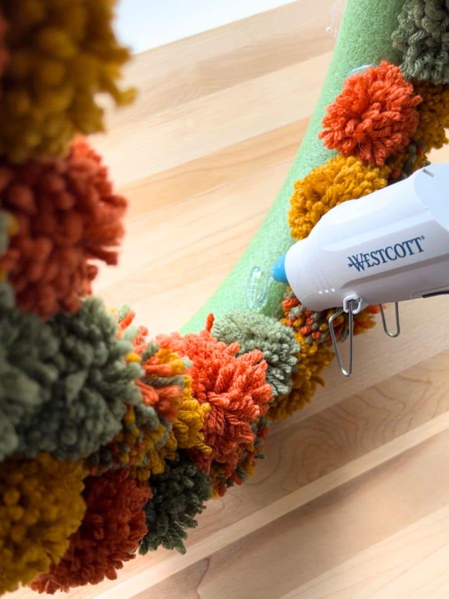 close up of glue gun putting dollop of glue on side of pompom covered wreath form