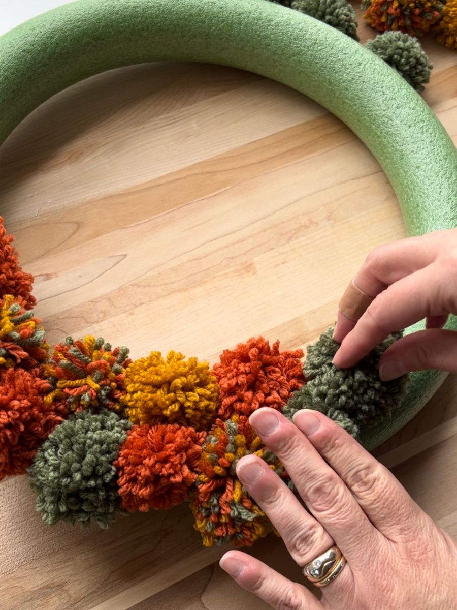 fingers pressing green pompom onto glue on foam wreath form