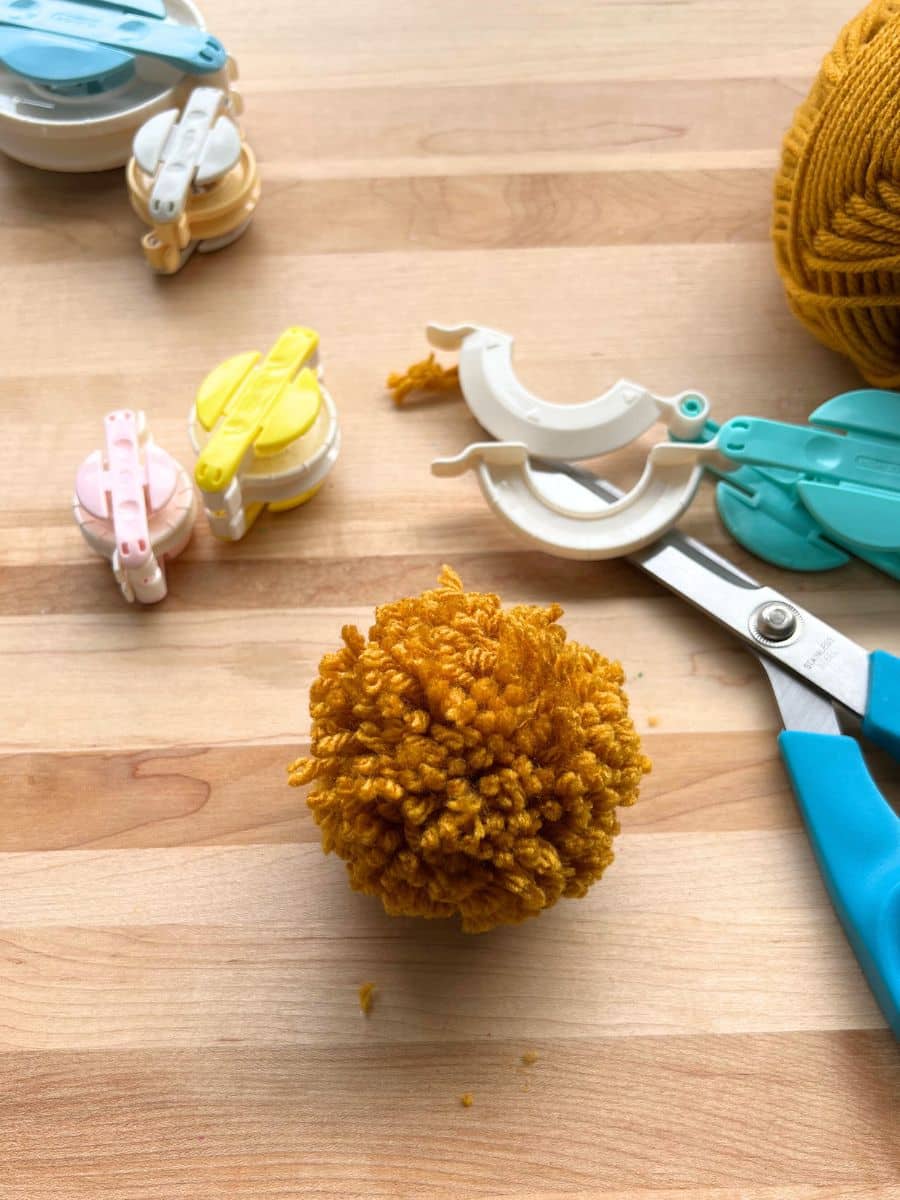 single brown pompom on table surrounded by scissors, yarn and pompom maker tool