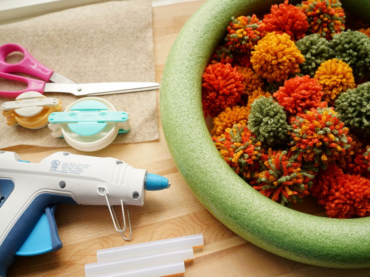 supplies for making pompom wreath - pom poms, green wreath form, scissors, glue gun, pom pom makers