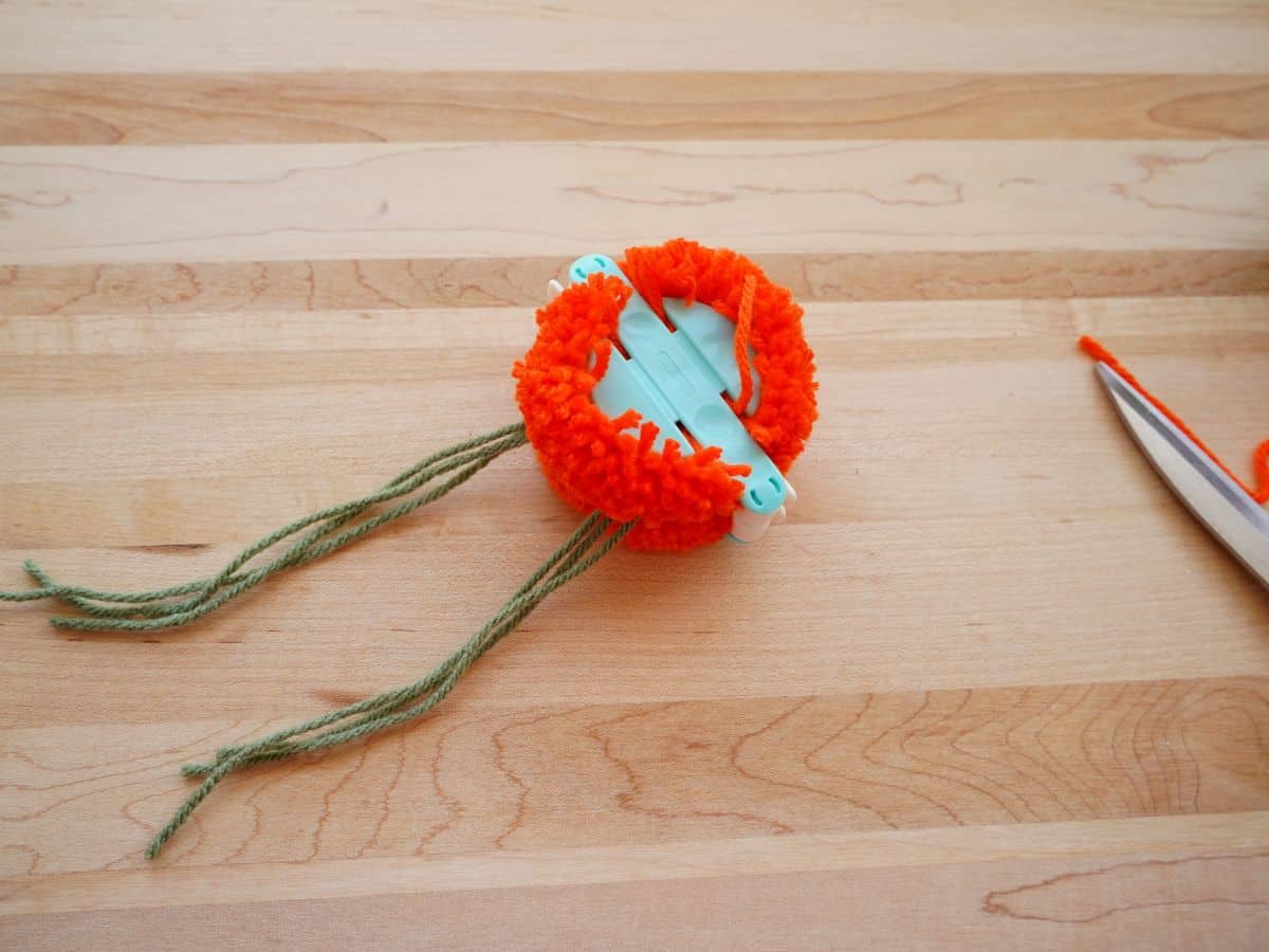 orange yarn around pom pom maker with green yarn wrapped around and knotted for pompom pumpkin