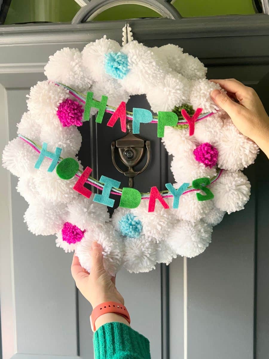 final christmas white pompom wreath hanging on front door, with happy holidays garland