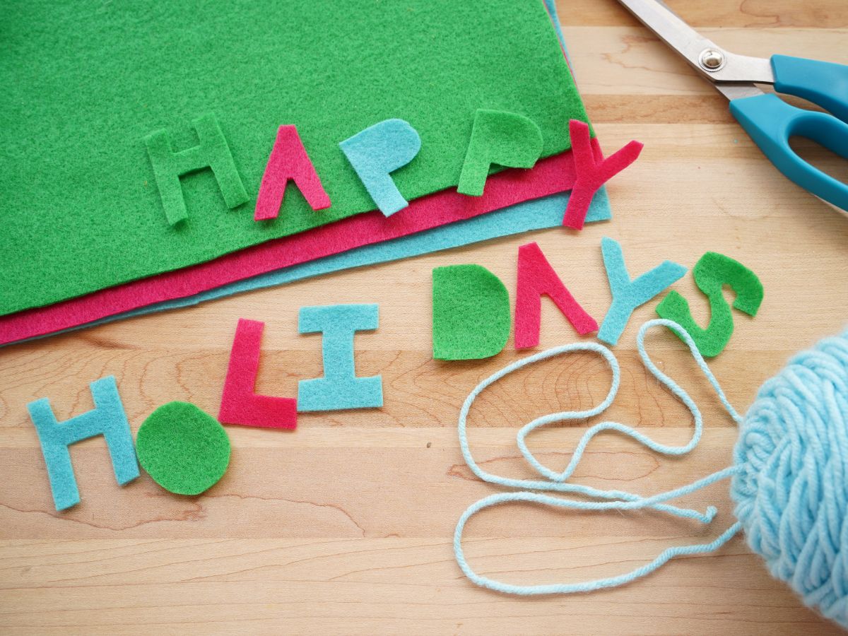 happy holidays letters cut out of green, pink and blue felt with scissors and blue yarn on table