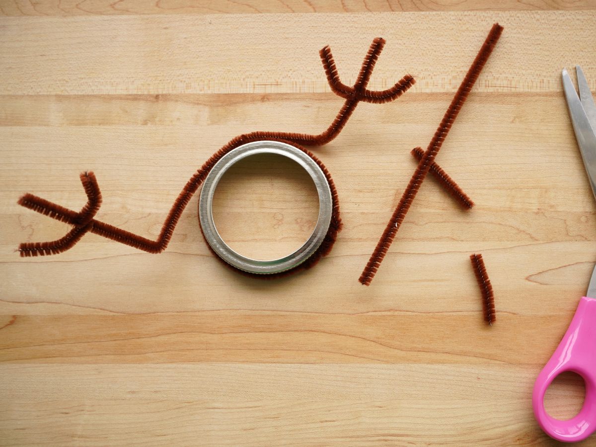 two brown pipe cleaners wrapped around mason jar lid to make antlers with pair of scissors beside