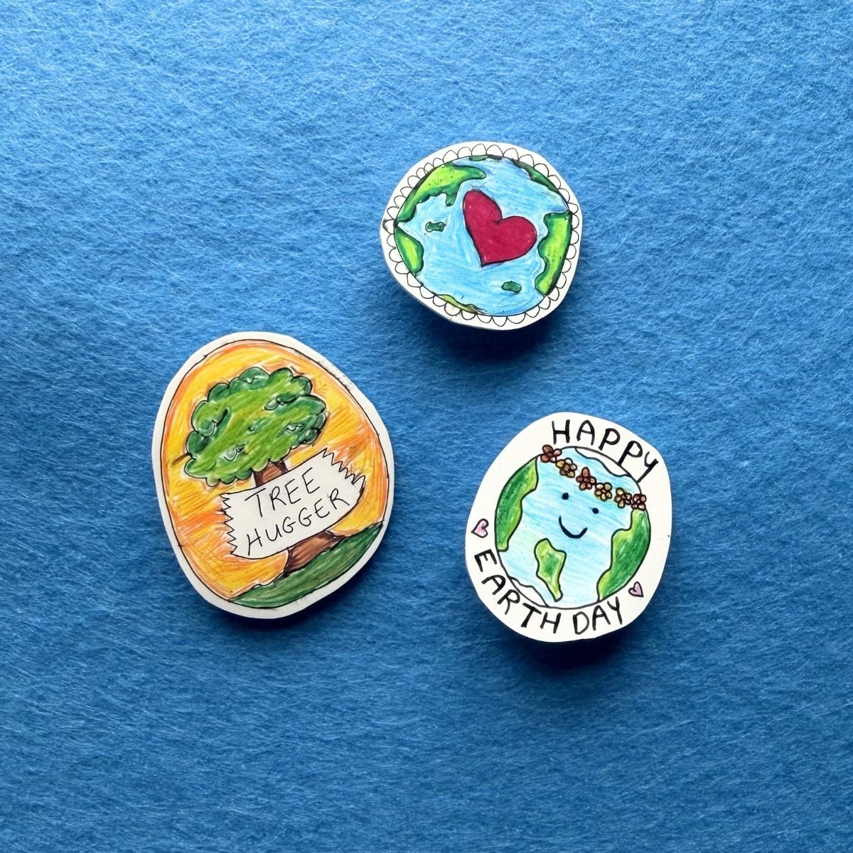 blue background with 3 homemade earth day pins - tree hugger pin and happy earth day pin and heart on earth pin