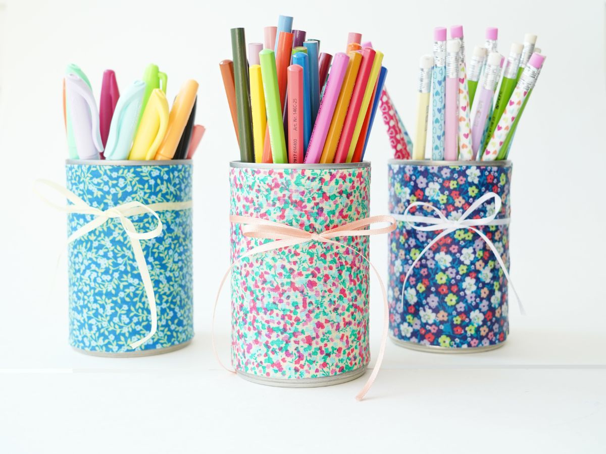 3 diy tin can pencil holders covered in fabric and wrapped with ribbons and filled with pencil crayons and markers