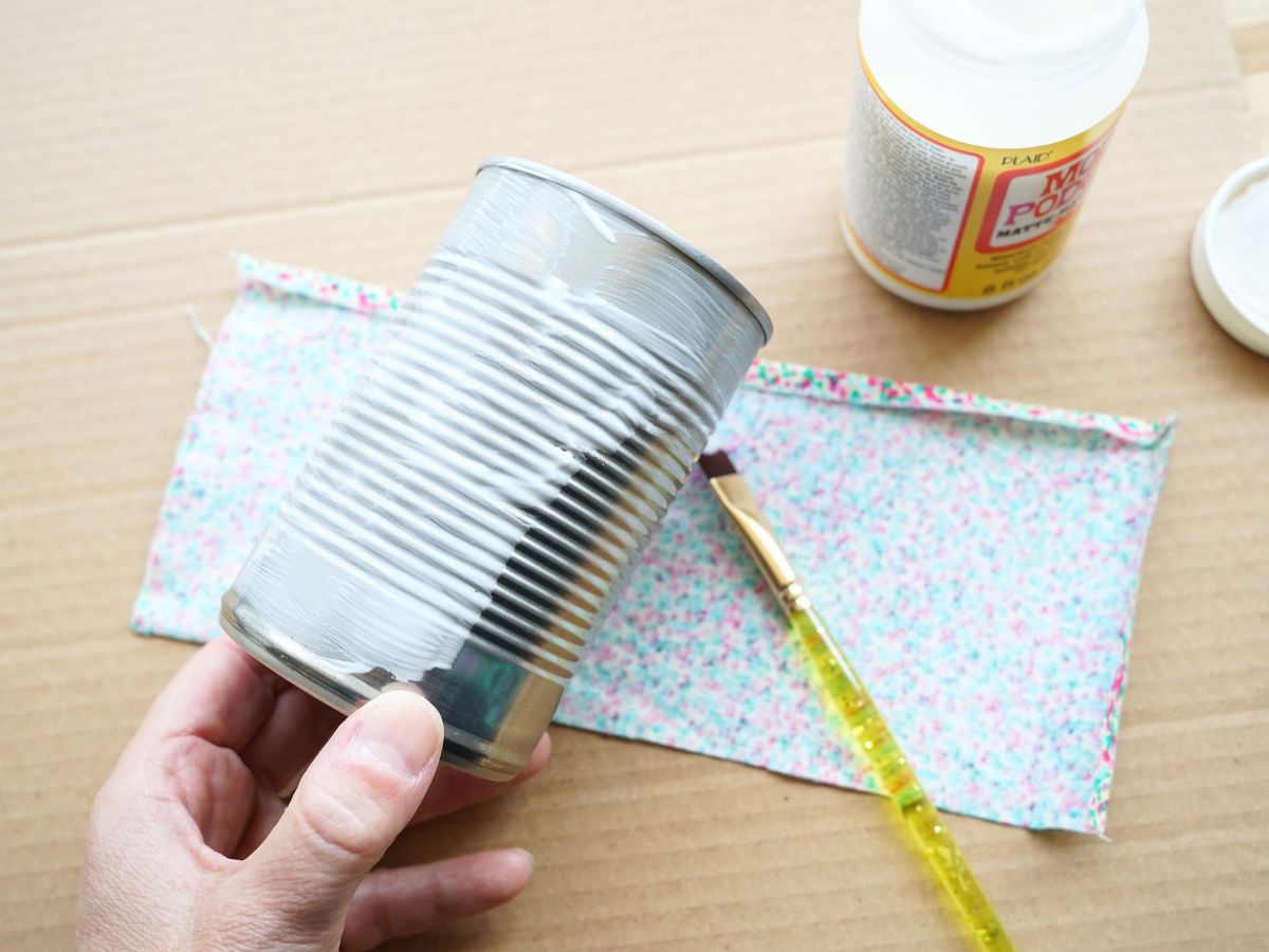 mod podge painted on tin can with paintbrush and piece of fabric laying on table below
