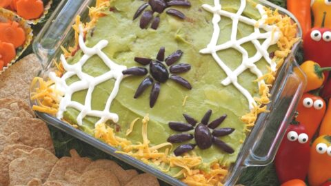 olive spider topped 7-layer halloween dip in glass pan surrounded by veggies and pumpkin tortilla chips