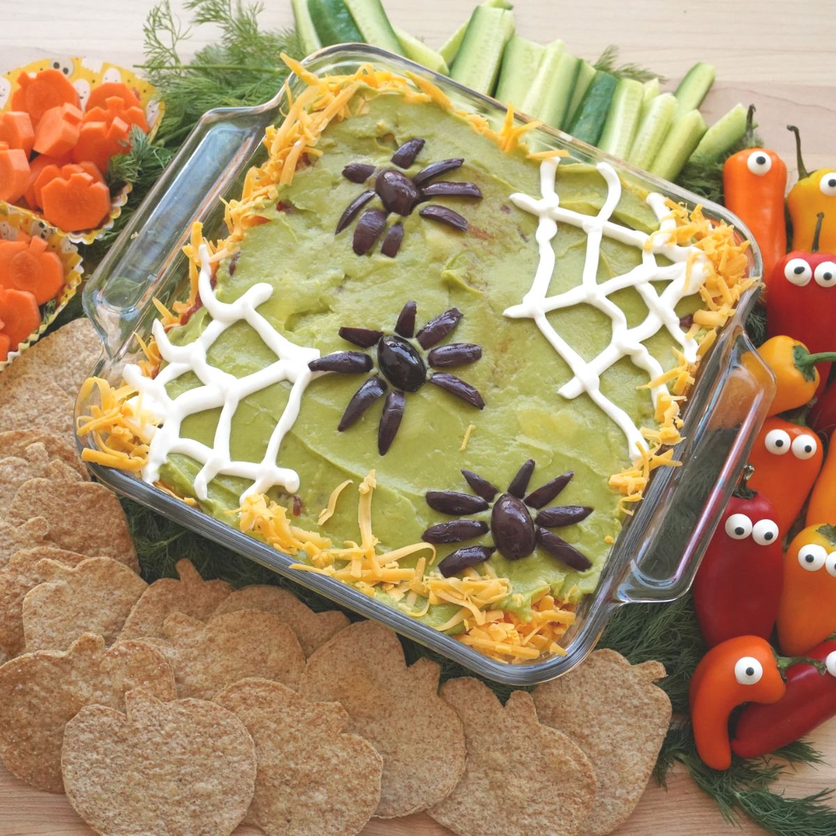 olive spider topped 7-layer halloween dip in glass pan surrounded by veggies and pumpkin tortilla chips