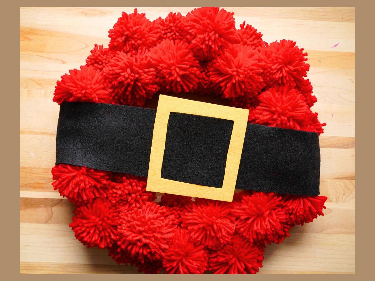 Red pom pom Santa wreath with Santa's belt sitting on table