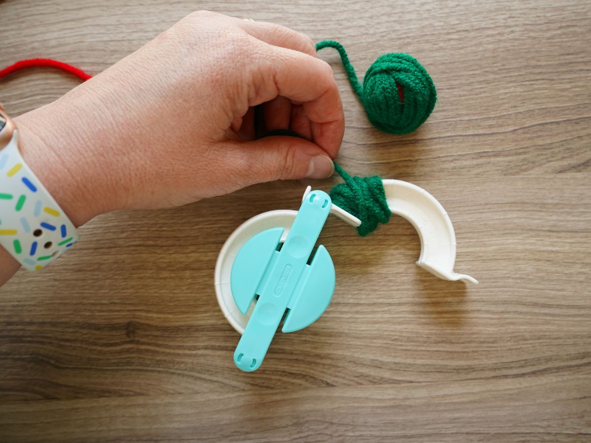 green yarn wrapped around half inch of one end of pom pom maker tool with woman's hand