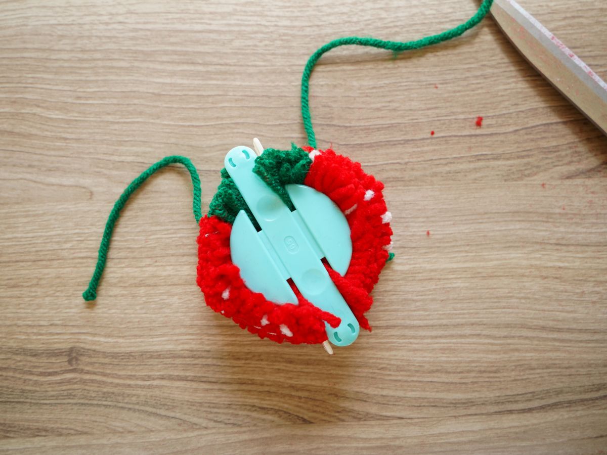 pom pom maker wrapped and cut and tied with green length of yarn for strawberry