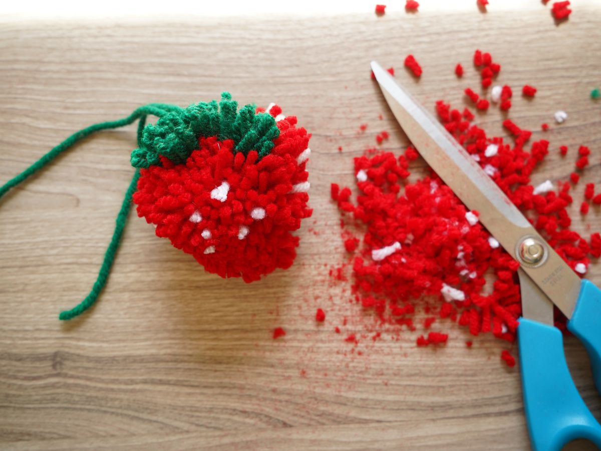 strawberry pom pom being trimmed into shape with scraps and scissors