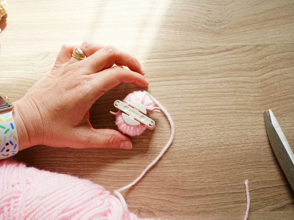 woman holding pompom maker tool that is wrapped with light pink yarn and a pair of scissors