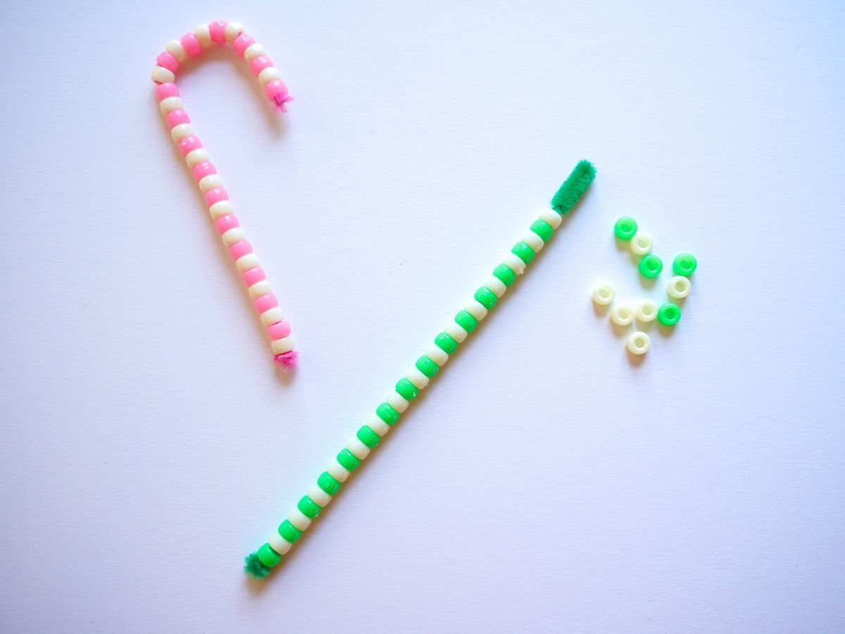 pink and white beaded candy cane beside a green pipe cleaner candy cane stick being beaded