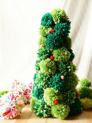 DIY pom pom Christmas tree standing on table surrounded by multi color pom poms featured image