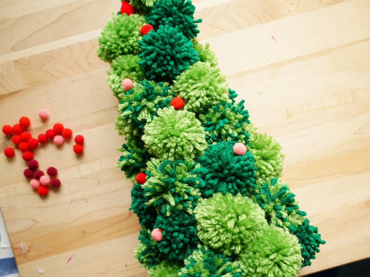 small red and pink pom poms being glued onto green pom pom christmas tree