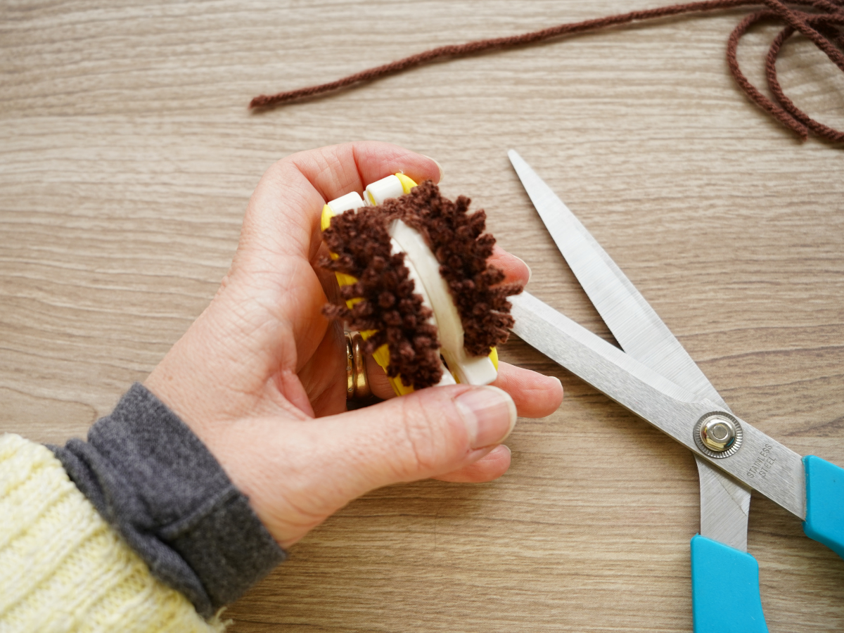 woman holding brown yarn wrapped pom pom maker being trimmed with scissors