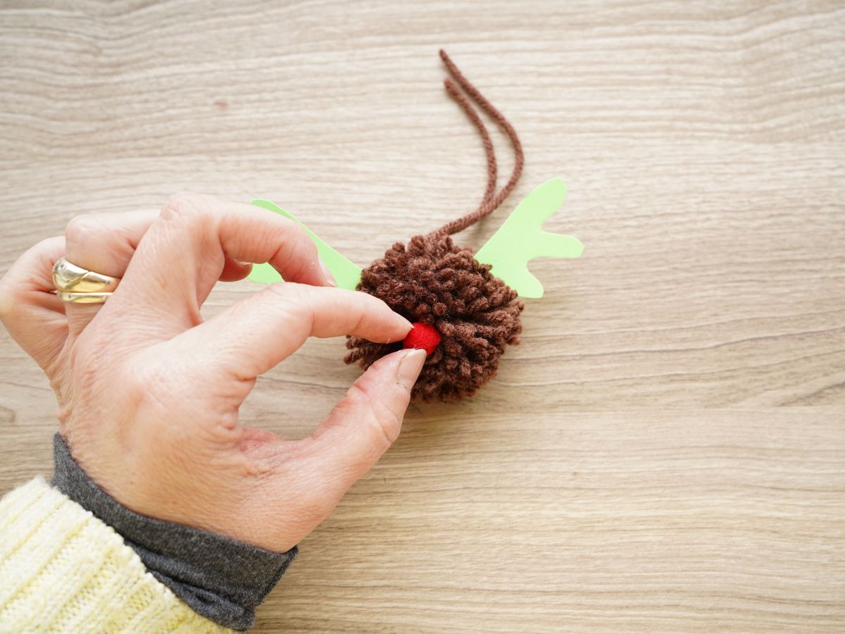 woman's hand pressing small red pom pom into nose of pom pom Rudolph ornament