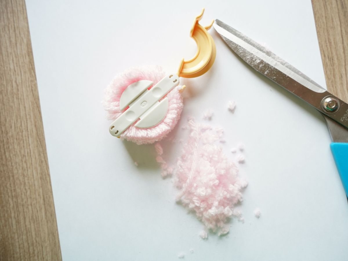 pom pom maker open and pink yarn being trimmed with scissors