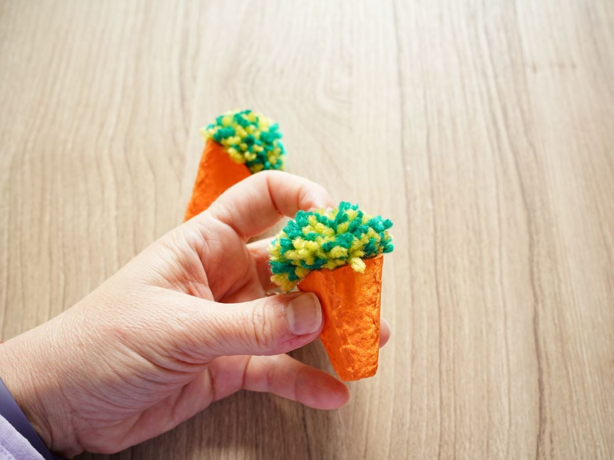 hand pressing green carrot top pom pom into the top of an egg carton carrot