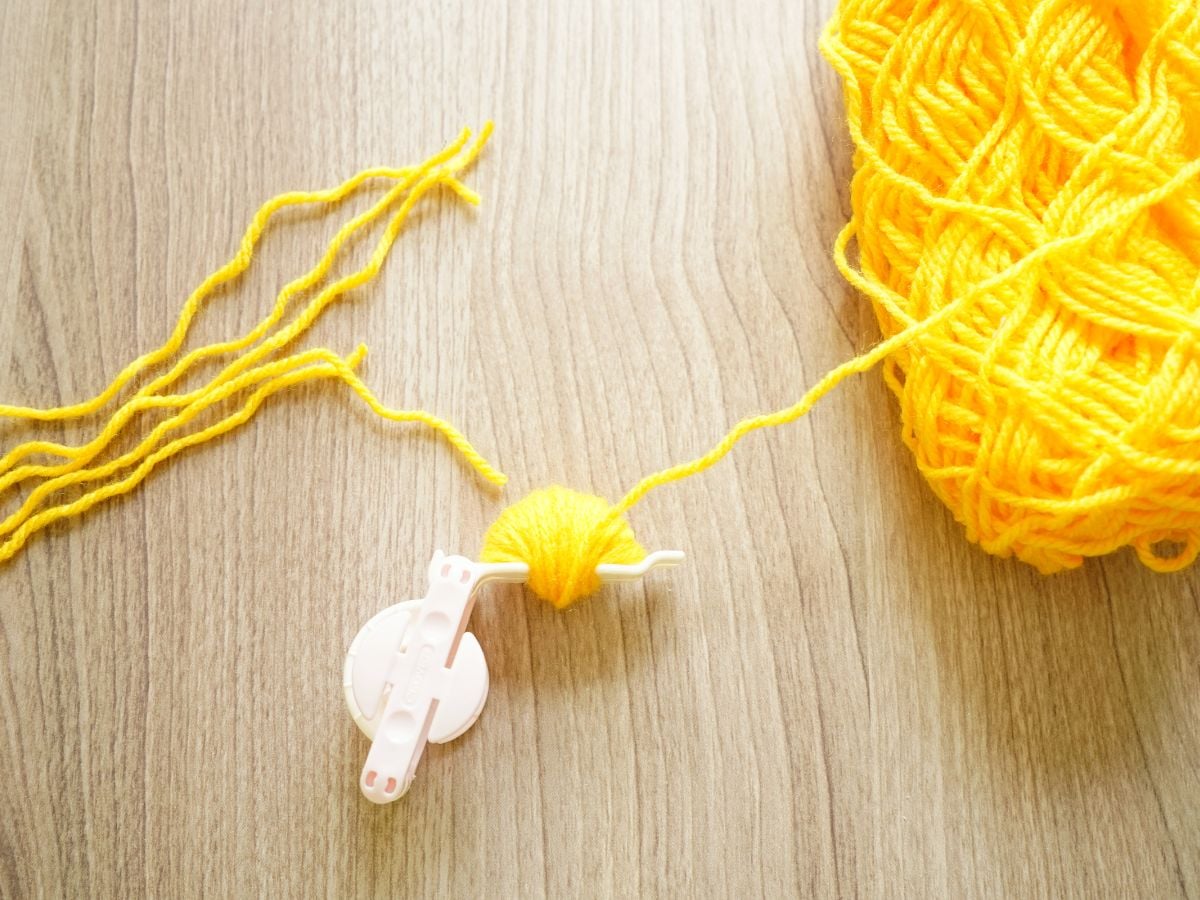 ball of yellow yarn beside a pom pom maker wrapped with yellow yarn on one half