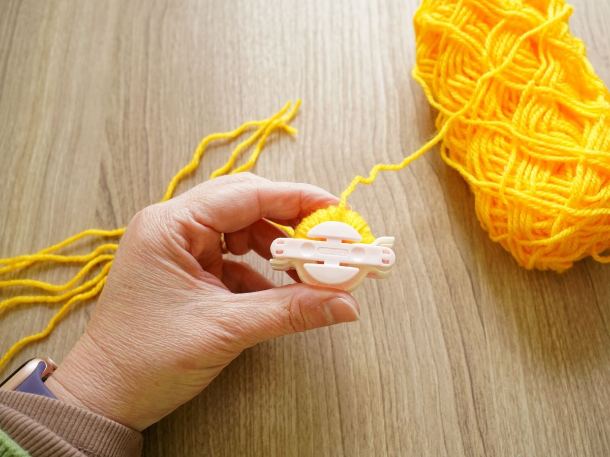 hand holding half wrapped pom pom maker that is wrapped in yellow thin yarn
